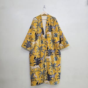 May include: A yellow, long-sleeved kimono-style robe with a vibrant jungle-themed pattern. The design features zebras, leopards, toucans, and palm trees. The robe is hanging on a wooden hanger against a plain white wall.