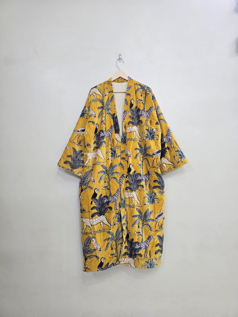May include: A yellow, long-sleeved kimono-style robe with a vibrant jungle-themed pattern. The design features zebras, leopards, toucans, and palm trees. The robe is hanging on a wooden hanger against a plain white wall.