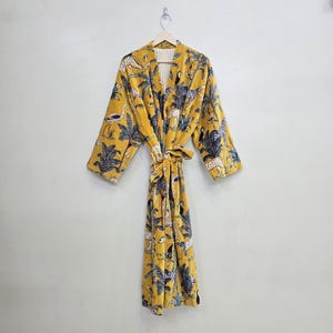 May include: A mustard yellow robe with a jungle animal print, including zebras, leopards, and birds. The robe has long sleeves and a matching tie belt. The interior lining is a cream color with vertical stripes. The robe is hanging on a wooden hanger.