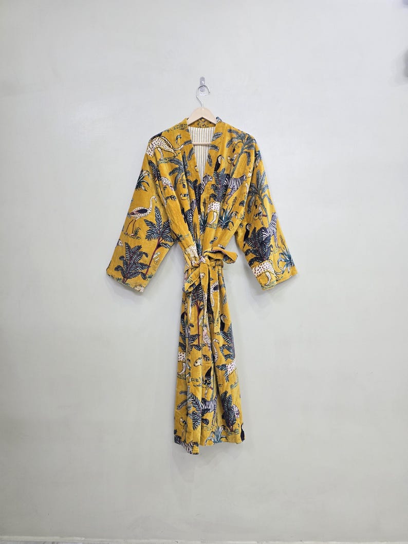 May include: A mustard yellow robe with a jungle animal print, including zebras, leopards, and birds. The robe has long sleeves and a matching tie belt. The interior lining is a cream color with vertical stripes. The robe is hanging on a wooden hanger.