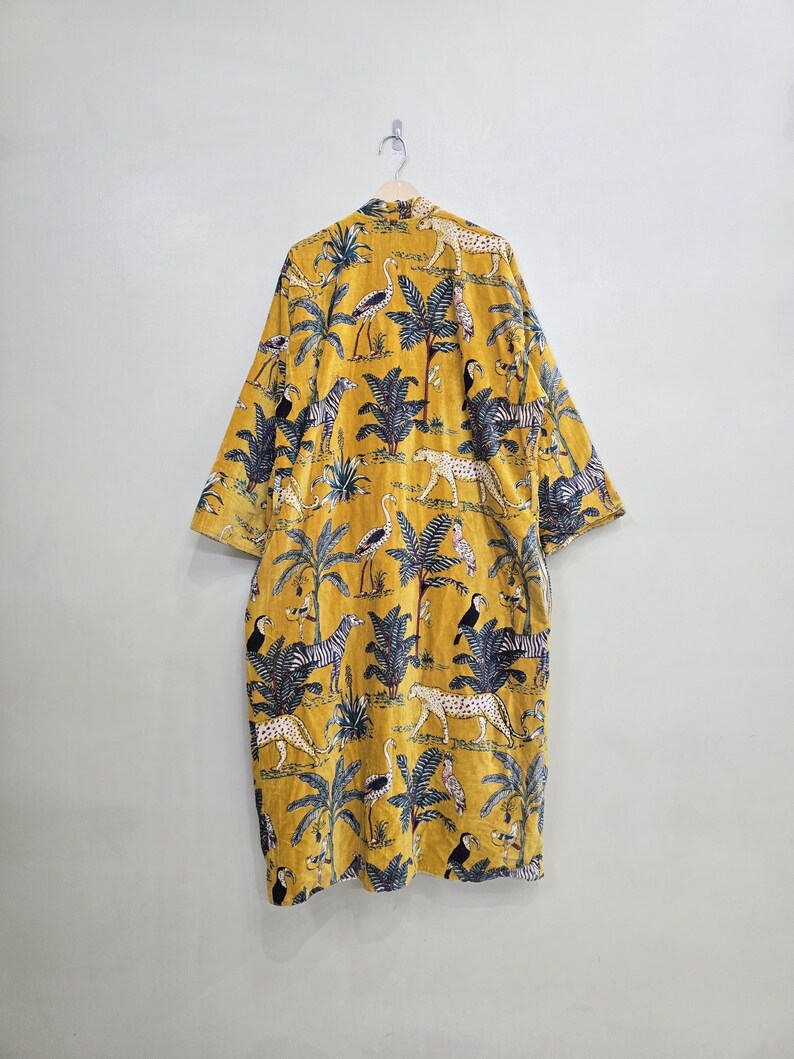 May include: Mustard yellow kimono-style robe with a jungle-themed pattern. The robe features a repeating design of zebras, leopards, toucans, and palm trees. The robe is hanging on a wooden hanger against a plain white wall.