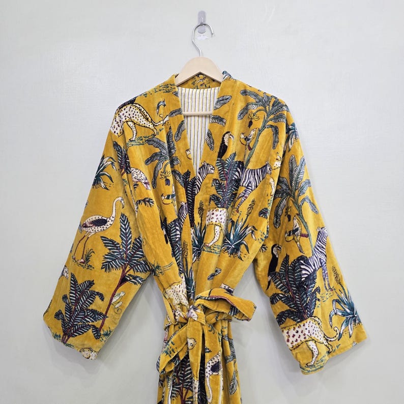 May include: Mustard yellow robe with a jungle animal print. The robe features a tie closure and a pattern of zebras, leopards, birds, and palm trees. The robe is hanging on a wooden hanger against a white wall.