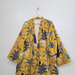 May include: Mustard yellow kimono-style robe with a jungle-themed pattern. The robe features zebras, leopards, toucans, and palm trees. The interior lining is striped. The robe is hanging on a wooden hanger.