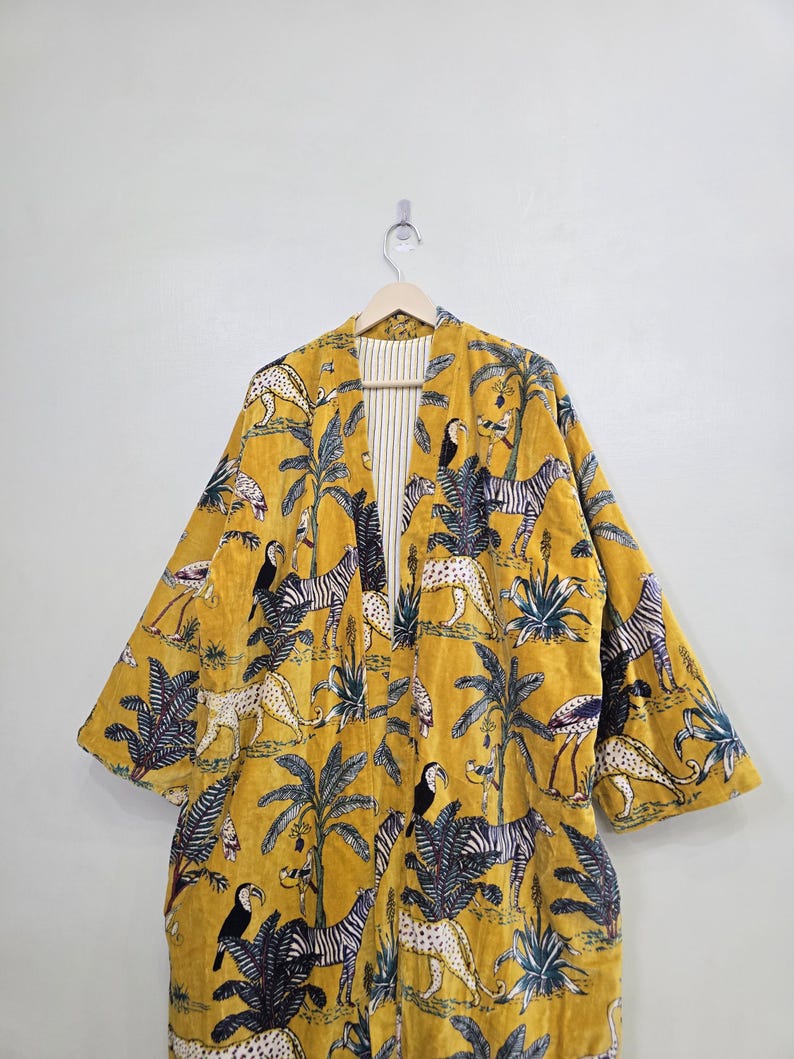 May include: Mustard yellow kimono-style robe with a jungle-themed pattern. The robe features zebras, leopards, toucans, and palm trees. The interior lining is striped. The robe is hanging on a wooden hanger.