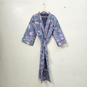 May include: A blue quilted robe with a floral pattern in pink and white hangs on a wooden hanger. The robe has long sleeves and a tie belt. The interior lining is a dark blue with a small white dot pattern.