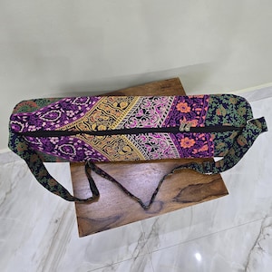 Może przedstawiać: Kolorowa torba na matę do jogi z zapięciem na zamek błyskawiczny i paskiem na ramię. Torba ma żywy wzór w odcieniach fioletu, pomarańczy i żółci, z czarnym zamkiem i paskiem. Torba ma około 71 cm długości.