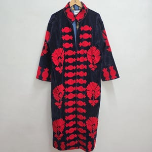 May include: A long, dark blue coat with vibrant red floral and candy-shaped embroidered designs. The coat has a high collar and long sleeves. The coat is hanging on a wooden hanger against a plain white background.