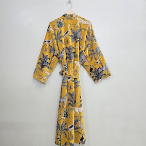 May include: Mustard yellow robe with a jungle-themed print. The robe features a repeating pattern of palm trees, zebras, leopards, and birds. It has long sleeves and a matching belt. The robe is hanging on a wooden hanger against a white wall.