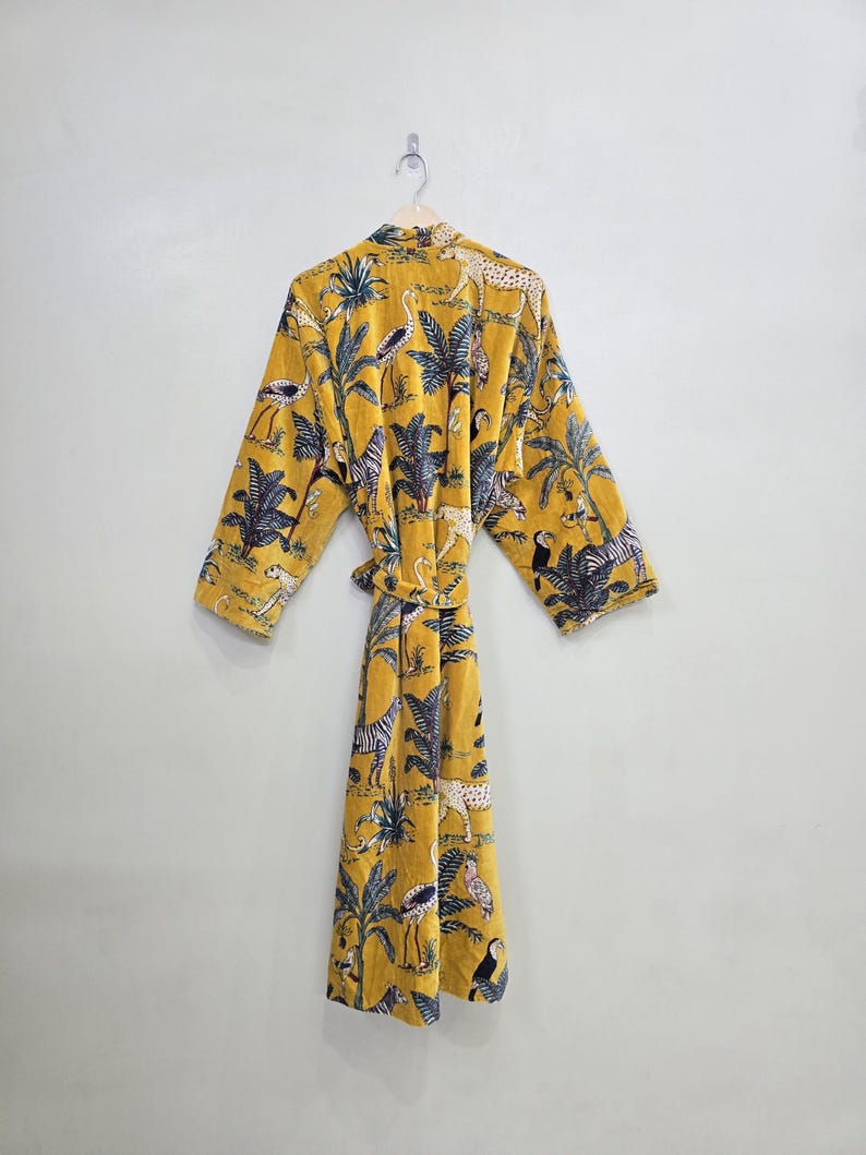 May include: Mustard yellow robe with a jungle-themed print. The robe features a repeating pattern of palm trees, zebras, leopards, and birds. It has long sleeves and a matching belt. The robe is hanging on a wooden hanger against a white wall.