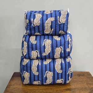 May include: Three stacked, rectangular, blue cosmetic bags with a vertical striped pattern. Each bag features a repeating leopard print in cream. The bags are stacked on a wooden surface.
