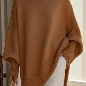 May include: A tan-coloured sweater with a relaxed fit. The sweater features a crew neck, long sleeves with ribbed cuffs, and a slightly asymmetrical hemline. The model is wearing white trousers and holding a tan handbag.