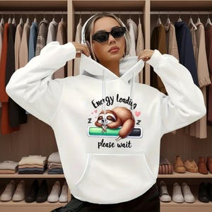 May include: White hoodie with a front pocket and a drawstring hood. The graphic on the front features a sleeping sloth with the text "Energy Loading, please wait". The sloth is brown and white, with a green loading bar.