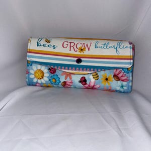 May include: A rectangular fabric wallet featuring a flap with horizontal yellow, pink, and blue stripes and the text "bees GROW butterflies". The lower section displays a light blue background with a colorful pattern of daisies, ladybugs, and bees.