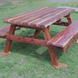 May include: A wooden picnic table with attached benches. The table and benches are made of reddish-brown wood. The table is rectangular with a slatted top. The benches are attached to the table's frame.