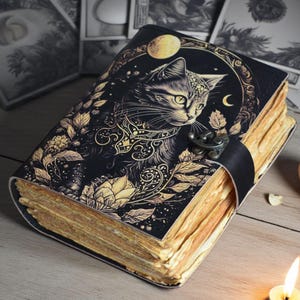 Puede incluir: Un diario negro con una ilustración de gato y detalles florales dorados. La portada presenta un dibujo detallado de un gato con una luna y estrellas. Las páginas tienen un aspecto vintage y envejecido, y una correa de cuero negro asegura el diario.
