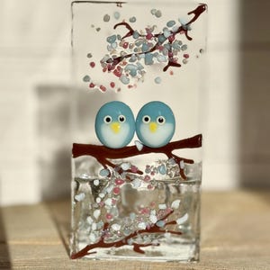 May include: A clear glass block featuring two blue bird ornaments on a brown branch. The glass is adorned with a branch design and colourful accents. The birds have white bellies and yellow beaks.