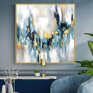 以下が含まれることがあります： 金色のフレームに入った抽象芸術絵画。このアートワークは、青、白、黒、金の筆致を組み合わせています。絵画は青い壁に飾られており、手前に青い椅子とネイビーブルーのキャビネットがあります。