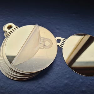 Könnte beinhalten: Ein Stapel goldfarbener, runder Ornamente mit einer abziehbaren Schutzschicht. Jedes Ornament hat oben eine kleine Schlaufe zum Aufhängen. Die Ornamente sind reflektierend und scheinen aus Metall zu sein.