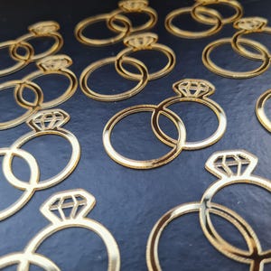 May include: A collection of gold-colored, reflective acrylic cutouts shaped like interlocking wedding rings with a diamond setting. Each piece features a circular band connected to a smaller ring and a diamond-shaped top with geometric lines.