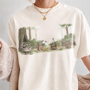 May include: Ivory-colored t-shirt featuring a forest scene with Grogu characters. The graphic shows Grogu in a pod and standing with other characters. The shirt is paired with pink and white checkered shorts. The text "COMFORT COLORS IVORY" is at the bottom.