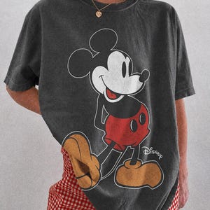 K&ouml;nnte beinhalten: Dunkelgraues T-Shirt mit einer Mickey Mouse-Grafik in Schwarz, Wei&szlig; und Rot. Auf dem T-Shirt ist das Wort "Disney" aufgedruckt. Der Text "COMFORT COLORS PEPPER" ist unten aufgedruckt. Teil der Comfort Colors-Kollektion.