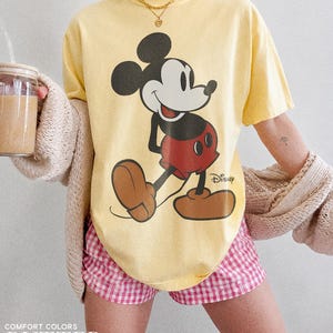 K&ouml;nnte beinhalten: Hellgelbes T-Shirt mit einem Mickey Mouse-Design in Schwarz, Rot und Braun. Das Shirt wird mit einer rosa-wei&szlig; karierten Shorts getragen. Die Worte "COMFORT COLORS" und "BUTTER" sind am unteren Rand des Bildes aufgedruckt.