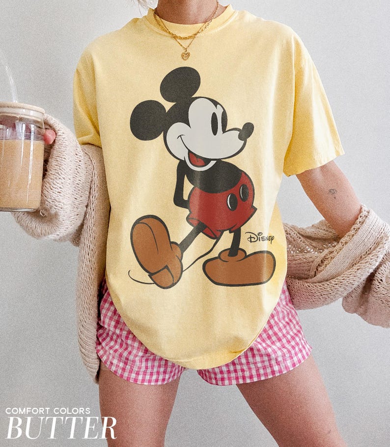 K&ouml;nnte beinhalten: Hellgelbes T-Shirt mit einem Mickey Mouse-Design in Schwarz, Rot und Braun. Das Shirt wird mit einer rosa-wei&szlig; karierten Shorts getragen. Die Worte "COMFORT COLORS" und "BUTTER" sind am unteren Rand des Bildes aufgedruckt.