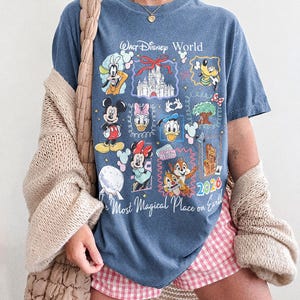 May include: Blue jean colored t-shirt featuring a colorful graphic of Disney characters and the text "Walt Disney World" and "The Most Magical Place on Earth". The shirt also has the year "2026" printed on it.