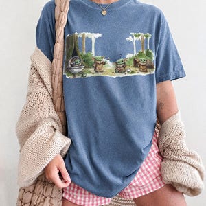 May include: Blue jean-colored t-shirt featuring a graphic of several Grogu characters in a forest setting. The shirt has the words "COMFORT COLORS" and "BLUE JEAN" printed at the bottom. The shirt is paired with pink and white checkered shorts.