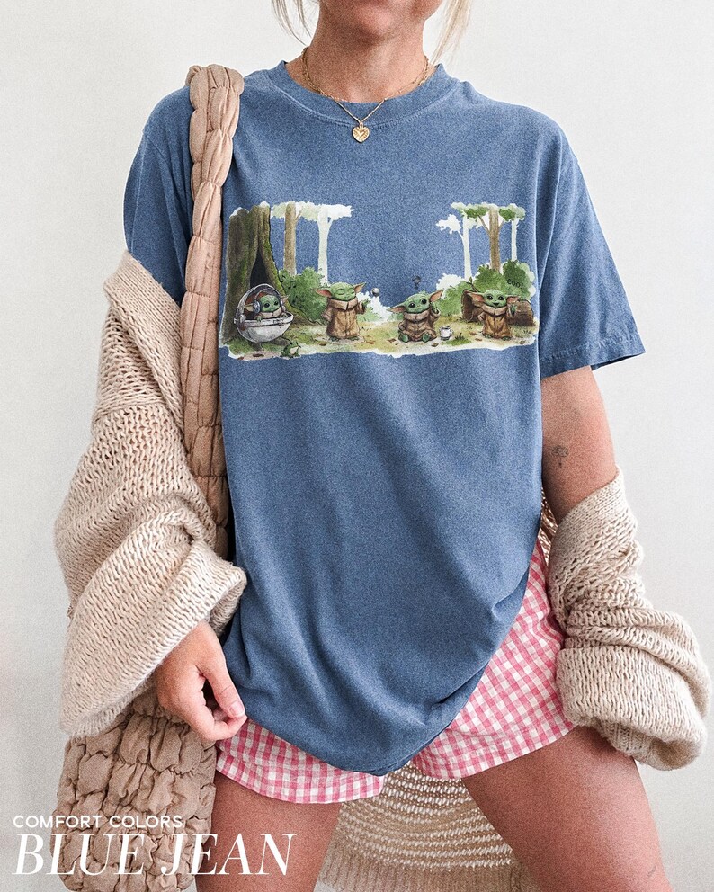 May include: Blue jean-colored t-shirt featuring a graphic of several Grogu characters in a forest setting. The shirt has the words "COMFORT COLORS" and "BLUE JEAN" printed at the bottom. The shirt is paired with pink and white checkered shorts.