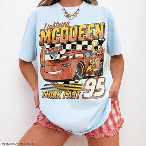 K&ouml;nnte beinhalten: Hellblaues T-Shirt mit einer Lightning McQueen-Grafik aus dem Film Cars. Das Design enth&auml;lt den Text "Lightning McQueen", "Cars", "95" und "Think Fast". Das Shirt ist ein Comfort Colors Chambray.