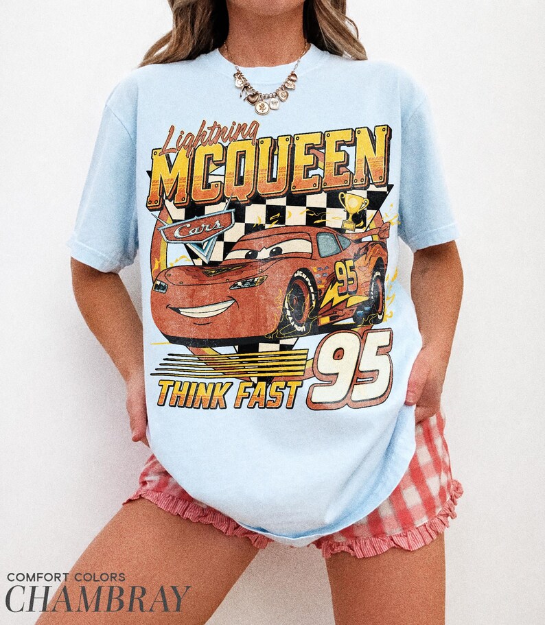 K&ouml;nnte beinhalten: Hellblaues T-Shirt mit einer Lightning McQueen-Grafik aus dem Film Cars. Das Design enth&auml;lt den Text "Lightning McQueen", "Cars", "95" und "Think Fast". Das Shirt ist ein Comfort Colors Chambray.