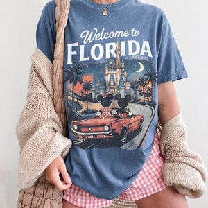 May include: Blue jean-colored t-shirt with a graphic that says "Welcome to Florida" and features a vintage car with Mickey and Minnie Mouse. The shirt also has a graphic of a castle, palm trees, and a crescent moon.