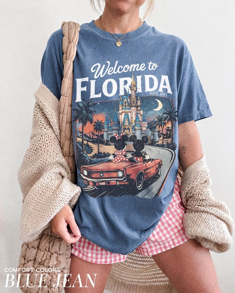May include: Blue jean-colored t-shirt with a graphic that says "Welcome to Florida" and features a vintage car with Mickey and Minnie Mouse. The shirt also has a graphic of a castle, palm trees, and a crescent moon.