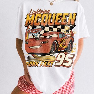 K&ouml;nnte beinhalten: Wei&szlig;es T-Shirt mit einem Aufdruck von Lightning McQueen aus dem Film Cars. Das Design enth&auml;lt die Texte "Lightning McQueen", "Cars", "95" und "Think Fast". Das T-Shirt hat einen Vintage-Look.