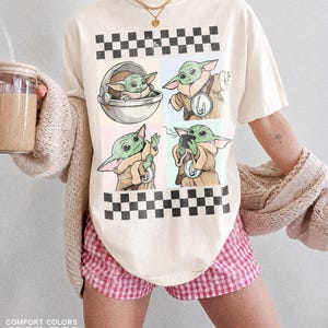 Puede incluir: Camiseta de color marfil con un dise&ntilde;o de cuatro paneles con im&aacute;genes de un personaje alien&iacute;gena verde. La parte superior e inferior de la camiseta tienen un patr&oacute;n de cuadros blancos y negros. El texto "COMFORT COLORS" e "IVORY" est&aacute; impreso en la parte inferior.