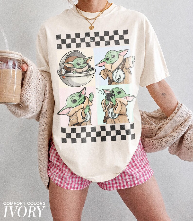 Puede incluir: Camiseta de color marfil con un dise&ntilde;o de cuatro paneles con im&aacute;genes de un personaje alien&iacute;gena verde. La parte superior e inferior de la camiseta tienen un patr&oacute;n de cuadros blancos y negros. El texto "COMFORT COLORS" e "IVORY" est&aacute; impreso en la parte inferior.