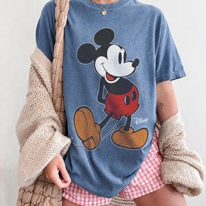 K&ouml;nnte beinhalten: Blaues T-Shirt mit einem Mickey Mouse-Motiv in Schwarz, Wei&szlig;, Rot und Beige. Das Wort "Disney" ist auf dem T-Shirt aufgedruckt. Der Text "COMFORT COLORS" und "BLUE JEAN" sind unten aufgedruckt. Das T-Shirt hat die Farbe Blue Jean.