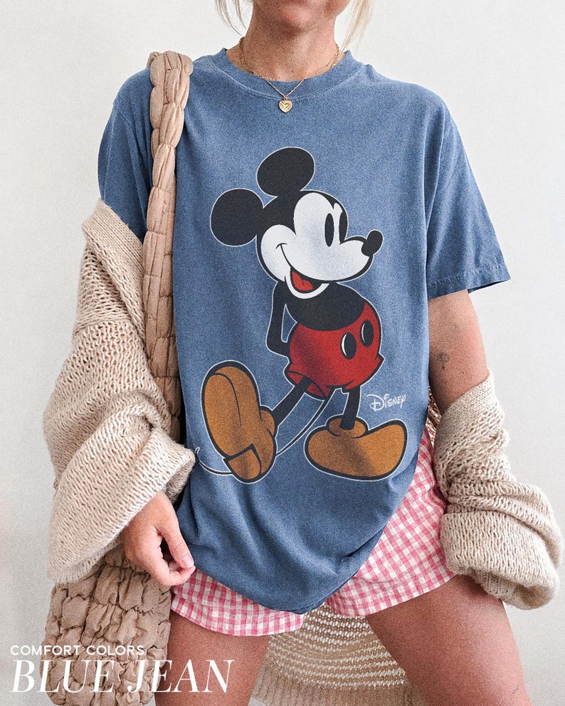 K&ouml;nnte beinhalten: Blaues T-Shirt mit einem Mickey Mouse-Motiv in Schwarz, Wei&szlig;, Rot und Beige. Das Wort "Disney" ist auf dem T-Shirt aufgedruckt. Der Text "COMFORT COLORS" und "BLUE JEAN" sind unten aufgedruckt. Das T-Shirt hat die Farbe Blue Jean.
