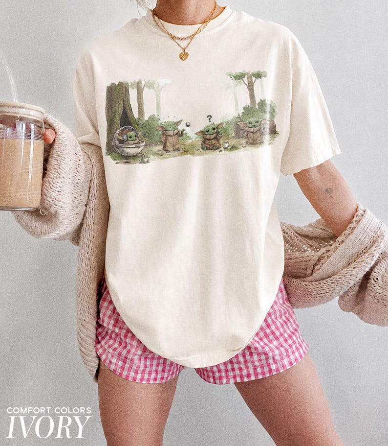 May include: Ivory-colored t-shirt featuring a forest scene with Grogu characters. The graphic shows Grogu in a pod and standing with other characters. The shirt is paired with pink and white checkered shorts. The text "COMFORT COLORS IVORY" is at the bottom.
