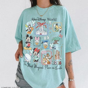 May include: A light blue t-shirt with a graphic featuring various Disney characters and the text "Walt Disney World" and "The Most Magical Place on Earth 2026". The shirt is labeled "Comfort Colors" and "Charky Mint".