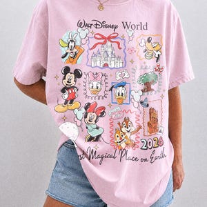 May include: A light pink t-shirt with a graphic featuring various Disney characters and the text "Walt Disney World" and "2026". The shirt also has the text "Most Magical Place on Earth".