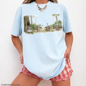 May include: Light blue t-shirt featuring a watercolor illustration of Grogu from The Mandalorian. The graphic shows Grogu in a pod and with other characters in a forest setting. The text "COMFORT COLORS CHAMBRAY" is printed at the bottom.