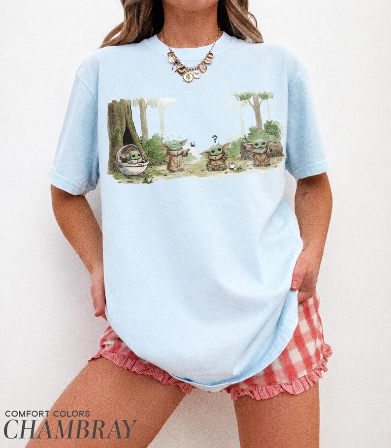 May include: Light blue t-shirt featuring a watercolor illustration of Grogu from The Mandalorian. The graphic shows Grogu in a pod and with other characters in a forest setting. The text "COMFORT COLORS CHAMBRAY" is printed at the bottom.