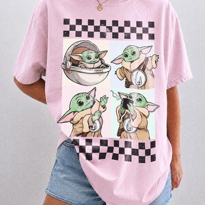 Puede incluir: Camiseta rosa claro con un dise&ntilde;o de cuatro paneles de un personaje alien&iacute;gena verde. El dise&ntilde;o incluye un patr&oacute;n de cuadros blanco y negro y la palabra "BLOSSOM" en texto blanco.