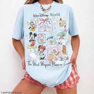 May include: Light blue t-shirt featuring Disney characters and the text "Walt Disney World" and "The Most Magical Place on Earth". The shirt has a 2026 graphic and is made of chambray fabric.