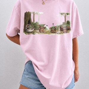 May include: A light pink t-shirt featuring a graphic of a character from a popular science fiction series. The graphic shows the character in various poses in a forest setting. The text "COMFORT COLORS BLOSSOM" is printed at the bottom.