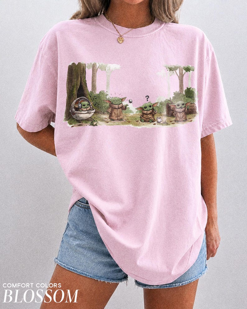 May include: A light pink t-shirt featuring a graphic of a character from a popular science fiction series. The graphic shows the character in various poses in a forest setting. The text "COMFORT COLORS BLOSSOM" is printed at the bottom.