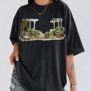 May include: Black t-shirt featuring a graphic of a character from a popular science fiction series. The design shows the character in a forest setting. The words "COMFORT COLORS BLACK" are printed at the bottom of the shirt.