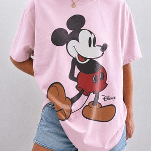 K&ouml;nnte beinhalten: Ein hellrosa T-Shirt mit einer gro&szlig;en Mickey Mouse-Grafik in Schwarz, Rot und Braun. Das Disney-Logo ist unter der Figur aufgedruckt. Das Shirt ist mit "Comfort Colors Blossom" beschriftet.